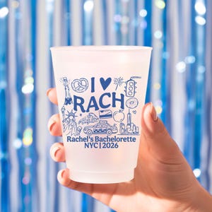 May include: A frosted plastic cup featuring blue ink graphics including a pretzel, heart, traffic light, donut, Statue of Liberty, pizza slice, taxi, and apple. The text reads I HEART RACH, Rachel's Bachelorette, and NYC 2026.
