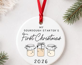 Puede incluir: Un adorno redondo de cerámica blanca que cuelga de una cinta roja. El texto dice MY SOURDOUGH STARTER'S First Christmas 2026. Debajo del texto hay tres ilustraciones de líneas de frascos de vidrio con masa madre, cada uno con una cubierta de tela.