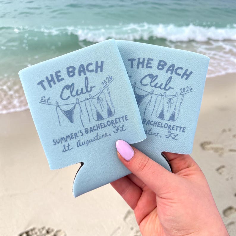 May include: Two light blue foam can coolers featuring dark blue text that reads THE BACH CLUB Est. 2026, SUMMER'S BACHELORETTE, St. Augustine, FL, and a graphic illustration of two bikini tops hanging on a clothesline.