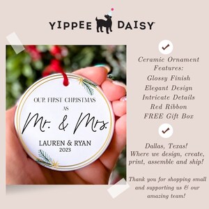 Op de afbeelding: Witte kerstbal van keramiek met gouden rand en groene dennentakken. De kerstbal heeft de tekst "Our First Christmas as Mr. & Mrs. Lauren & Ryan 2023".