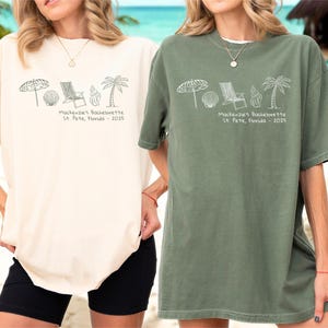 May include: Two oversized t-shirts, one cream and one olive green, with a beach-themed graphic. The graphic includes an umbrella, seashell, beach chair, and palm tree. Text on the shirts reads "Mackenzie's Bachelorette St. Pete, Florida - 2025."