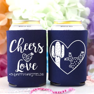 May include: Two navy blue can coolers with white text and designs. One cooler says "Cheers to Love #OHHAPPYHARTFIELDS". The other cooler has a heart with a cactus and the text "T+J".