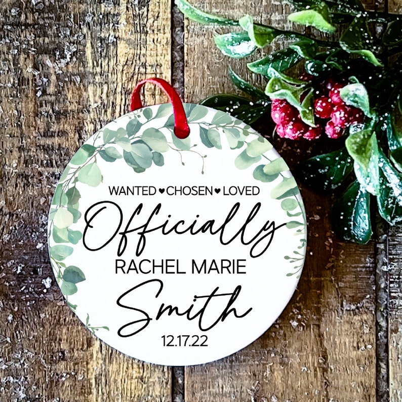Op de afbeelding: Ronde witte ornament met een groen bladerenontwerp. De ornament heeft zwarte tekst die "WANTED CHOSEN LOVED Officially Rachel Marie Smith 12.17.22" luidt.
