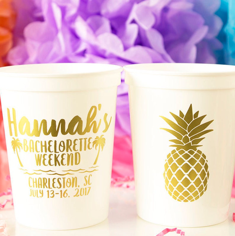 May include: Two white plastic cups with gold lettering and designs. One cup says "Hannah's Bachelorette Weekend Charleston, SC July 13-16, 2017". The other cup has a gold pineapple design.