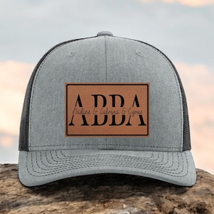 May include: A gray trucker hat with a black mesh back. The front features a brown rectangular patch with the name "ADDA" in bold black letters above "Nadine & Sabrina & Cyrus" and "ADDA" below.