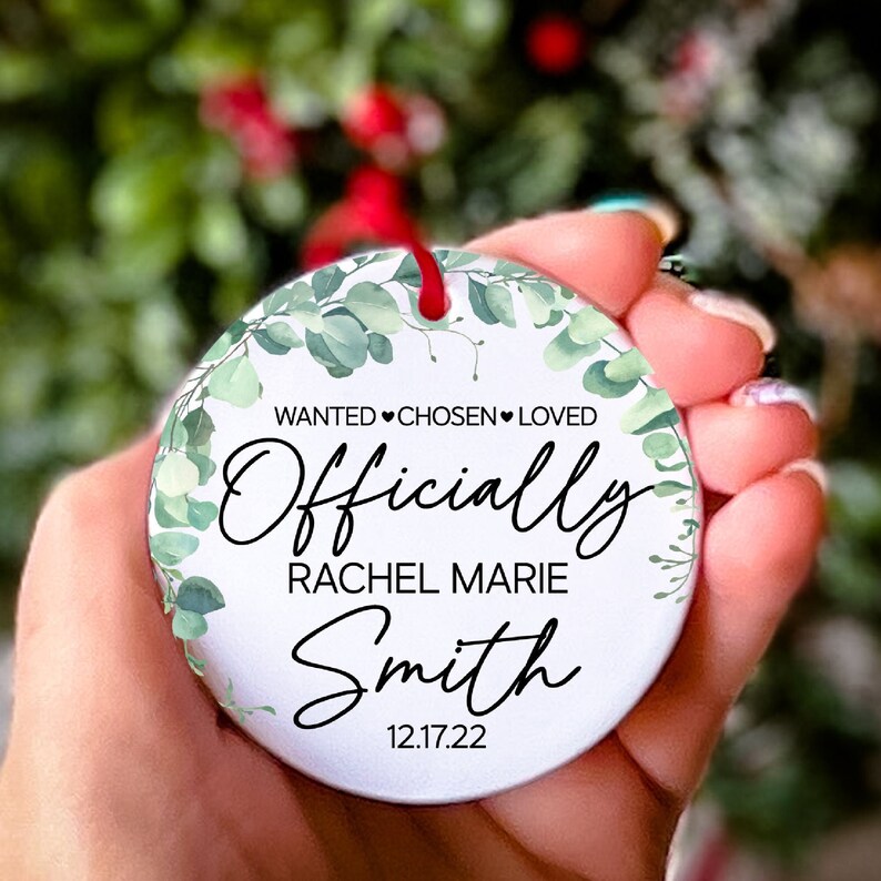 Op de afbeelding: Ronde witte ornament met groene aquarelbladeren en de tekst "WANTED CHOSEN LOVED Officially Rachel Marie Smith 12.17.22"