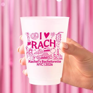 May include: A white plastic cup featuring hot pink graphics including a pretzel, a heart, a traffic light, the Statue of Liberty, a slice of pizza, a taxi, an apple, and the text I HEART RACH, Rachel's Bachelorette NYC 2026.