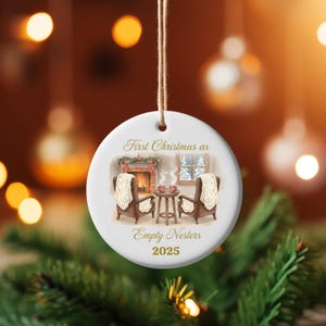 Op de afbeelding: Een witte keramische kerstbal met een aquarel illustratie van een gezellige woonkamer scène. De ornament toont een open haard, twee fauteuils en de tekst "First Christmas as Empty Nesters 2025."