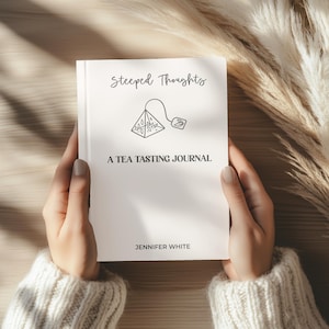 May include: A white journal titled "Steeped Thoughts" with the subtitle "A Tea Tasting Journal" and the author's name, Jennifer White. The cover features a line drawing of a tea bag. The journal is held by a person wearing a cream-colored sweater.