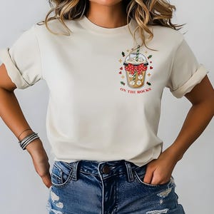Peut inclure: T-shirt crème avec un motif d'une boisson dans un gobelet transparent avec des glaçons, un nœud rouge et le texte "ON THE ROCKS". Le motif comprend des cœurs et des étoiles.