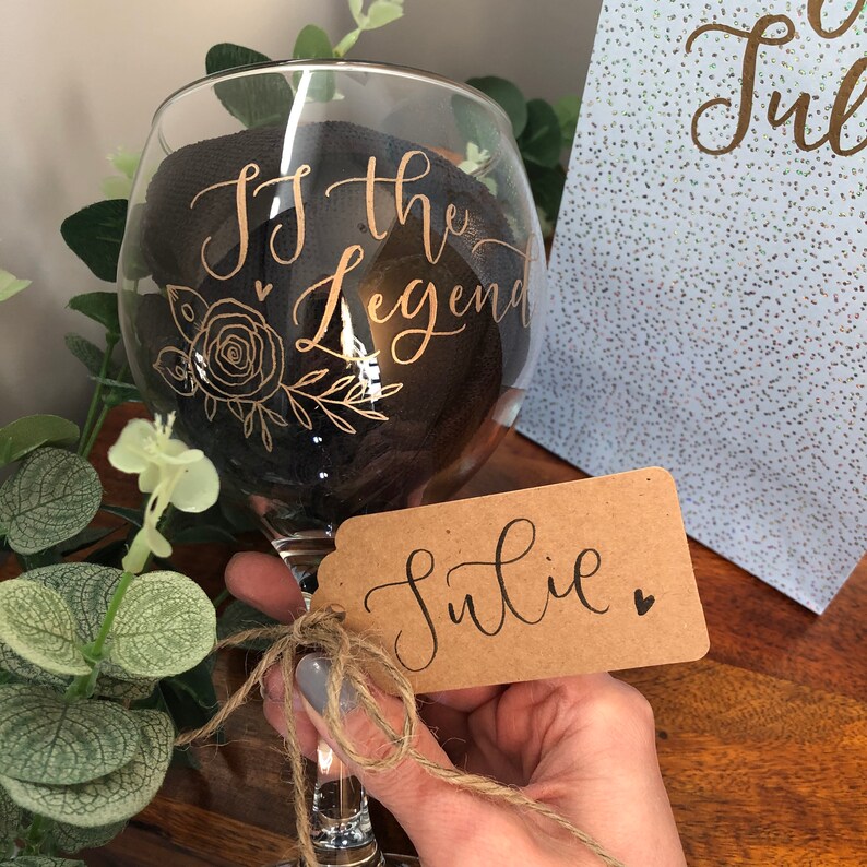 May include: A clear glass with a black ribbon and gold lettering that reads "To the Legend". The glass is decorated with a gold floral design. A brown gift tag with the name "Julie" written in black cursive is tied to the glass with twine.