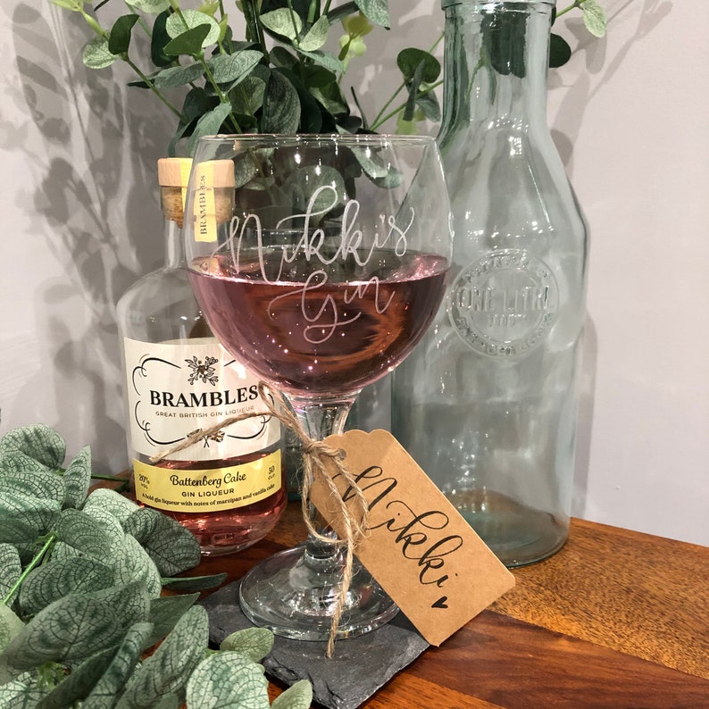 May include: A personalized gin glass with the text "Nikki's Gin" etched on the side. The glass is filled with a pink gin drink and is sitting on a black slate coaster. A brown gift tag with the text "Nikki" is tied to the glass with twine.