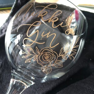 May include: A clear glass gin glass with the words "Nikki's Gin" etched in gold lettering. The glass also features a gold floral design with a rose.