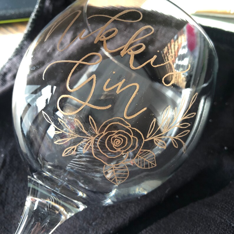 May include: A clear glass gin glass with the words "Nikki's Gin" etched in gold lettering. The glass also features a gold floral design with a rose.