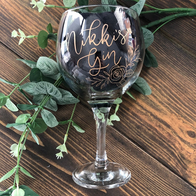 May include: A clear glass gin glass with a gold inscription that reads "Nikki's Gin" and a floral design.