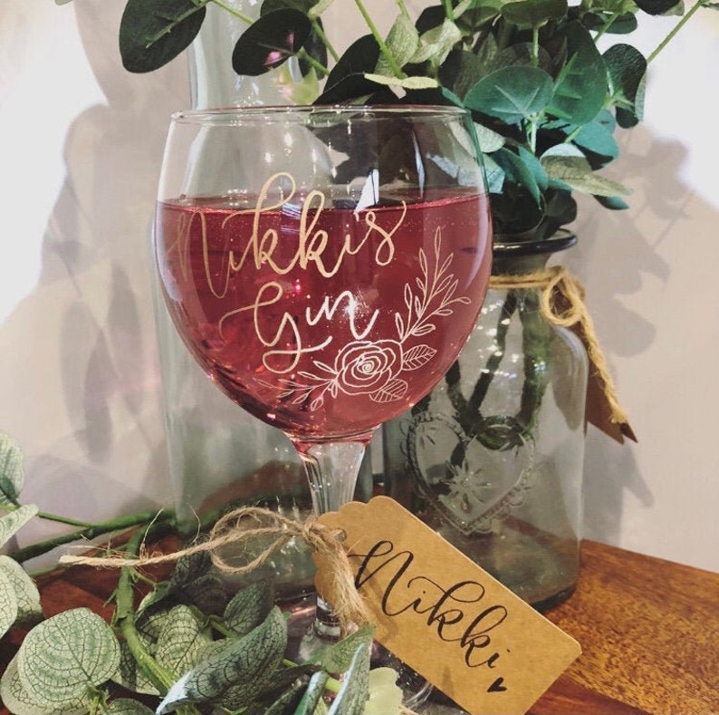 May include: A personalized gin glass with the text "Nikki's Gin" in gold lettering and a floral design. The glass is filled with a pink drink and has a brown tag with the text "Nikki" in gold lettering.