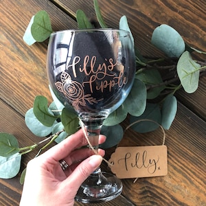 May include: A clear glass gin glass with a gold design that says "Filly's Tipple" and a floral design. A brown paper tag with the word "Filly" written in gold is attached to the glass.