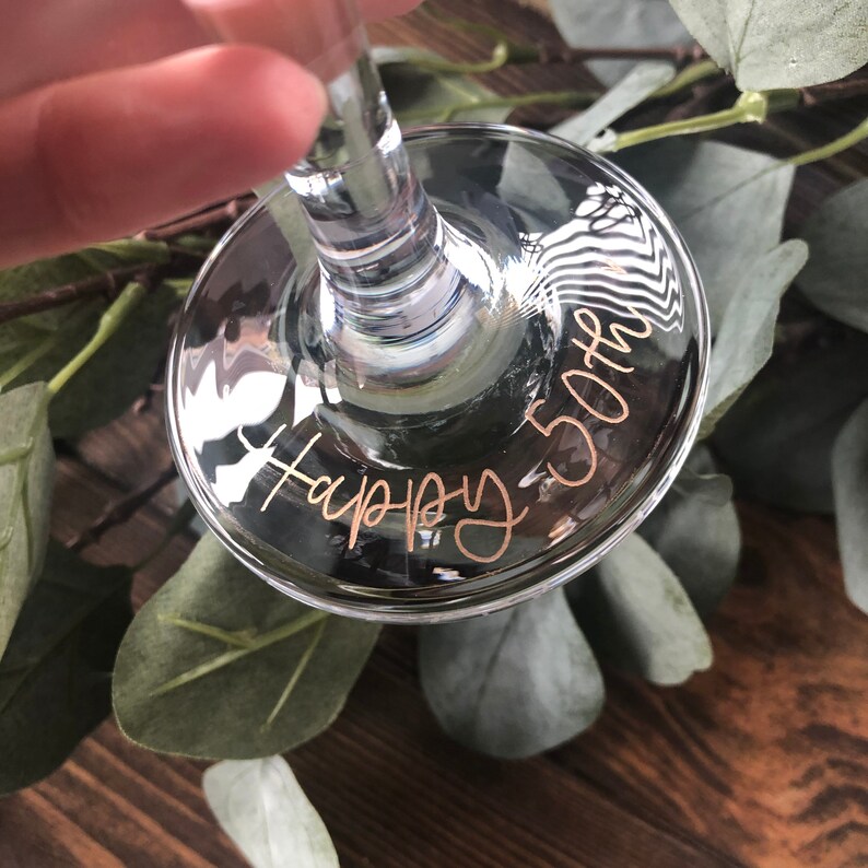 May include: A clear glass wine glass with the words "Happy 50th" etched on the bottom in gold lettering.