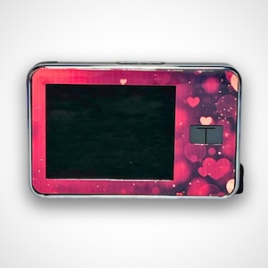 May include: A small, silver-colored electronic device with a red and pink heart-shaped pattern. The device has a small screen and a button on the front.