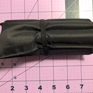 Puede incluir: Una bolsa negra enrollada con cierre de banda el&aacute;stica. La bolsa est&aacute; sobre una superficie rosa con cuadr&iacute;cula.
