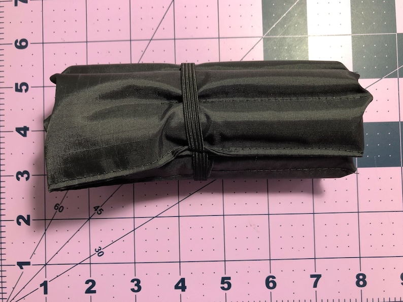 Puede incluir: Una bolsa negra enrollada con cierre de banda el&aacute;stica. La bolsa est&aacute; sobre una superficie rosa con cuadr&iacute;cula.