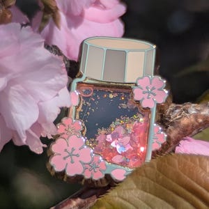 Puede incluir: Un pin decorativo de esmalte con forma de tarro, lleno de purpurina rosa y detalles de flores de cerezo. El pin tiene un borde azul claro y está adornado con flores de cerezo rosas. El fondo presenta flores rosas y una rama marrón.