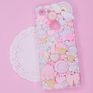 Peut inclure: Une coque de téléphone transparente remplie de breloques en résine rose pastel, bleu et blanc. Les breloques comprennent des cornets de glace, des étoiles, des cœurs et des nuages. La coque a un arc-en-ciel et un nuage sur le dessus.