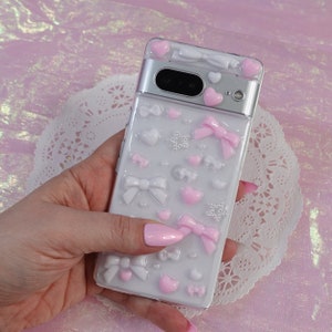 Könnte beinhalten: Eine weiße Handyhülle mit durchsichtiger Rückseite und rosa und weißen 3D-Verzierungen, darunter Schleifen, Herzen und Sterne. Die Hülle ist für ein Smartphone mit Kamerabuckel konzipiert.