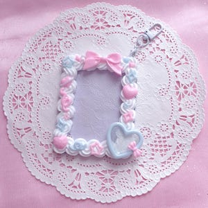 Può includere: Una cornice decorativa con una combinazione di colori bianco, rosa e azzurro. La cornice è adornata con cuori e fiocchi, e una chiusura in metallo. È posizionata su un centrino di pizzo bianco, su uno sfondo rosa.
