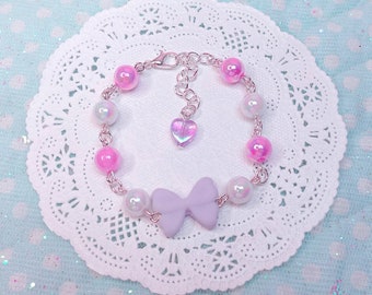 May include: A pastel pink and white beaded bracelet with a purple bow charm and a silver heart charm. The bracelet is on a white lace doily.