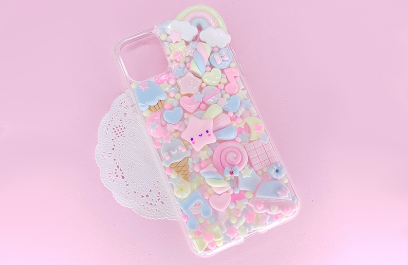 May include: A clear phone case with a pink, blue, and yellow pastel color scheme. The case is decorated with various 3D charms, including stars, hearts, candy, and a rainbow.