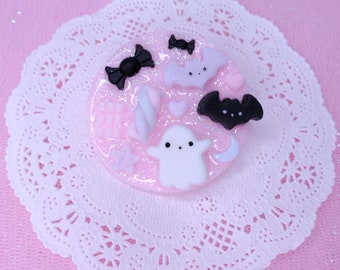 May include: A pink, circular Halloween-themed decoration with various miniature charms. The charms include a white ghost, black bats, a black bow, a pink heart, and a candy. The decoration is on a white lace doily.