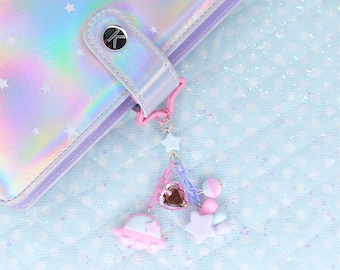 Puede incluir: Una carpeta de color púrpura iridiscente con un patrón de estrellas y un amuleto rosa y azul adjunto. El amuleto presenta un corazón, estrellas y una nube.
