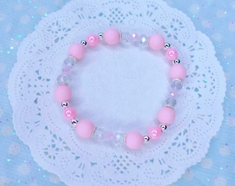May include: A pink and clear beaded bracelet with silver accents. The bracelet is made with round beads and is on a white lace doily.