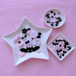 Può includere: Un piatto bianco a forma di stella con decorazioni in pasta di zucchero rosa e nera, tra cui un pipistrello, un coniglietto, un lecca-lecca e una nota musicale. Ci sono due piccole ciotole, una con codette nere e rosa e l'altra con perline nere e rosa.