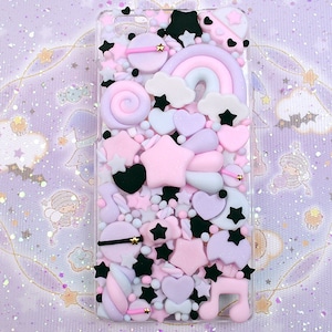 May include: A clear phone case filled with colorful, pastel-colored, 3D decorations. The decorations include stars, hearts, a rainbow, a lollipop, and a music note. The case is on a purple and white glittery background.