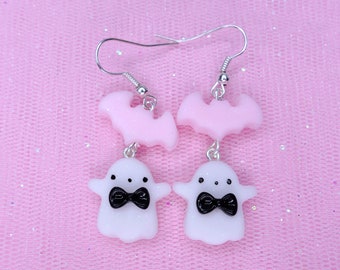 May include: A pair of dangle earrings featuring a pink bat charm and a white ghost charm with a black bow tie. The earrings are on a pink background.