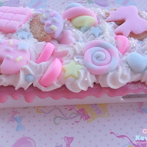 May include: A pink and white phone case decorated with various pastel-coloured candy and sprinkles. The case has a white background with a pink border and a white phone cutout. The case is decorated with a variety of candy, including lollipops, gumdrops, and chocolate bars. The case is also decorated with sprinkles and glitter.