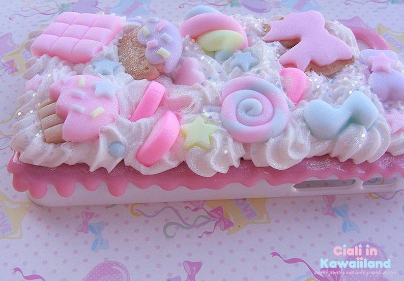 May include: A pink and white phone case decorated with various pastel-coloured candy and sprinkles. The case has a white background with a pink border and a white phone cutout. The case is decorated with a variety of candy, including lollipops, gumdrops, and chocolate bars. The case is also decorated with sprinkles and glitter.