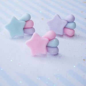 May include: Three pastel-colored star and cloud earrings. The earrings are made of polymer clay and are in shades of pink, blue, and purple.