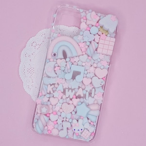 May include: A clear phone case with a pink, blue and white kawaii design. The case is filled with various charms, including stars, hearts, rainbows and a musical note. The text "KAWAII" is spelled out in pink and white letters.