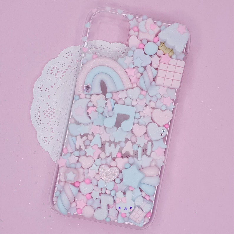 May include: A clear phone case with a pink, blue and white kawaii design. The case is filled with various charms, including stars, hearts, rainbows and a musical note. The text "KAWAII" is spelled out in pink and white letters.