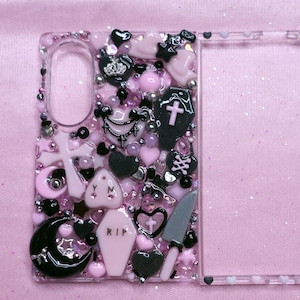 May include: A clear phone case decorated with a dense arrangement of pink and black charms, including a black coffin with a pink cross, a pink coffin with the text RIP, a grey knife, a black crescent moon, hearts, stars, and various beads.