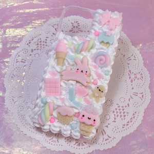 Puede incluir: Una funda transparente para teléfono decorada con diseños 3D de fondant en rosa pastel, azul y blanco. Los diseños incluyen un conejito, un cupcake, una piruleta, un bastón de caramelo y un caramelo en espiral.