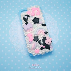 May include: A white phone case with a 3D design featuring a pink and blue rainbow, stars, a cupcake, a music note, and a black bunny with a crescent moon. The case is decorated with white frosting.