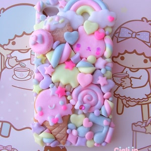 May include: A colorful phone case decorated with pastel-colored fondant-like decorations, including a rainbow, clouds, hearts, stars, a cookie, a lollipop, and an ice cream cone. The case is decorated with the text "COOKIE" and "Ciali in Kawaiiland".