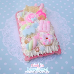 May include: A pink and white phone case decorated with colourful candy and sweets, including a doughnut, a lollipop, and marshmallows. The case is covered in glitter and has a white background.