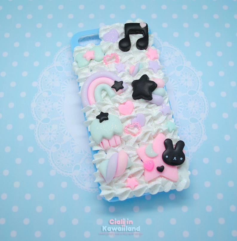 May include: A blue phone case decorated with white frosting, pink hearts, pastel rainbow, black stars, and a black bunny with a pink nose. The case has a glittery finish.