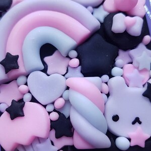 Op de afbeelding: Een close-up van een telefoonhoesje versierd met pastelkleurige 3D-charms. De charms omvatten een regenboog, een hart, een ster, een werveling en een konijn. De achtergrond is zwart.
