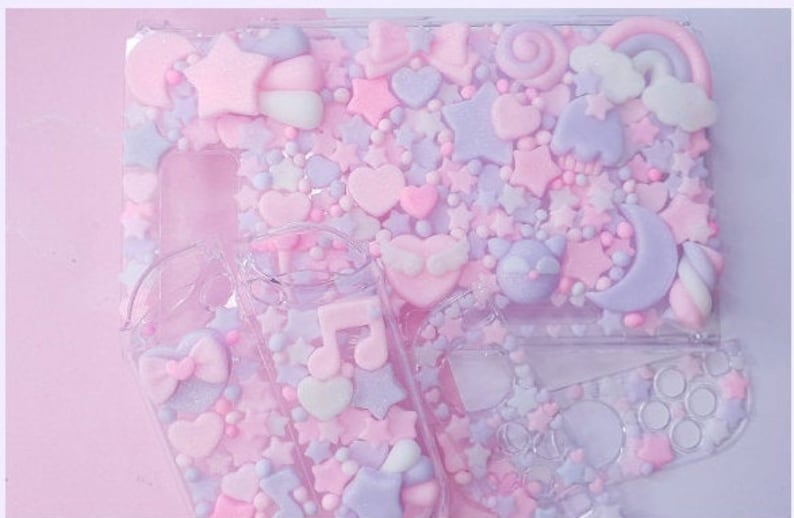 Puede incluir: Una funda de pl&aacute;stico transparente decorada con formas 3D en rosa pastel, morado y blanco, incluyendo estrellas, corazones y una luna creciente. La funda est&aacute; llena de peque&ntilde;as cuentas y purpurina de colores.